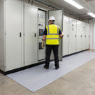 Electrician in hi-vis, safety helmet and insulating gloves standing on light grey broad-rib switchboard matting laid along the front of a row of low-voltage switchgear panels