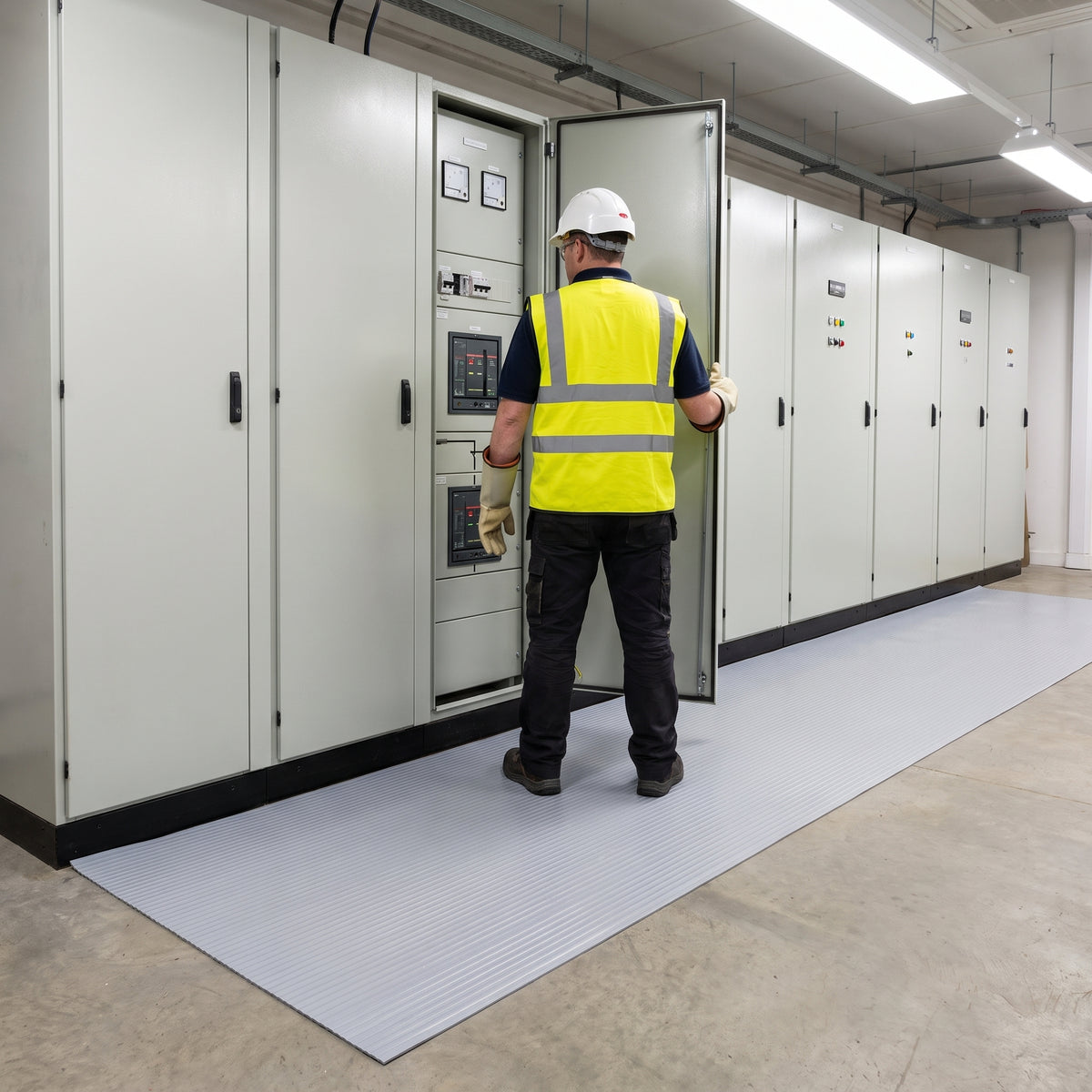Electrician in hi-vis, safety helmet and insulating gloves standing on light grey broad-rib switchboard matting laid along the front of a row of low-voltage switchgear panels