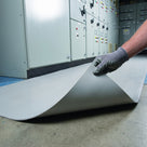 Close view of light grey broad-rib switchboard matting with a gloved hand lifting one corner to show the thin flexible 5mm section and smooth underside, switchgear behind