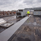 Grey open-lattice roof walkway matting loose laid across a dark EPDM membrane roof from the access hatch to the air-handling plant, engineer in hi-vis walking along it