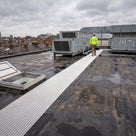 White open-lattice walkway matting across a wet black EPDM roof from the hatch to rooftop plant, engineer walking it