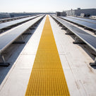 Yellow open-lattice walkway matting as a long straight run between rows of solar panels on a pale membrane roof