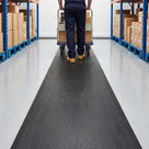 Black 14mm close-mesh PVC matting runner down a clean warehouse aisle, a worker pushing a loaded flatbed trolley along it