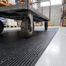 Close view of a flatbed trolley wheel rolling across black 14mm close-mesh matting, showing the fine ribs and mat thickness