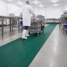 Green 14mm close-mesh PVC matting runner through a clean food production hall, an operative in white wheeling a stainless trolley of crates