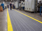 Grey ribbed foam matting with integral yellow safety edge marking a pedestrian walkway through a factory