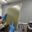 Blue-gloved hands holding an olive-green Tufnol 10G/40 glass epoxy laminate sheet in a workshop, glass weave visible on the face