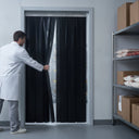 Worker in a white coat parting black PVC strip curtains at a cold store doorway, stocked shelving right