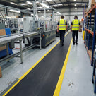 Black open mesh matting with bright yellow safety edging marking a pedestrian walkway along a factory aisle