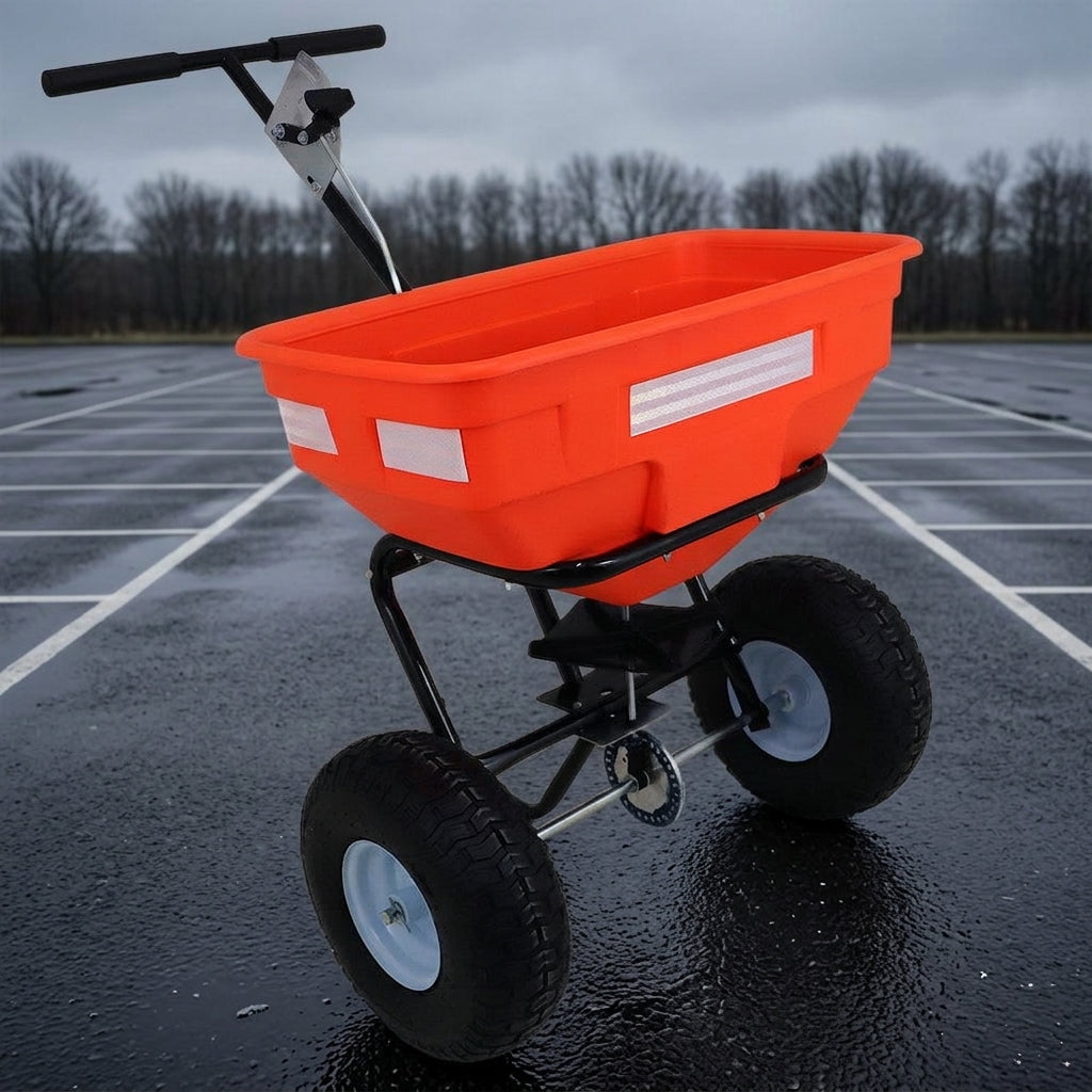 Orange walk-behind salt spreader with black pneumatic wheels standing on a wet marked-out car park under a grey winter sky