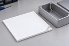 White silicone sponge square with its release liner peeled back at one corner, alloy enclosure and screwdriver behind
