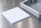 White silicone sponge square on a brushed steel bench, corner of an alloy enclosure behind it